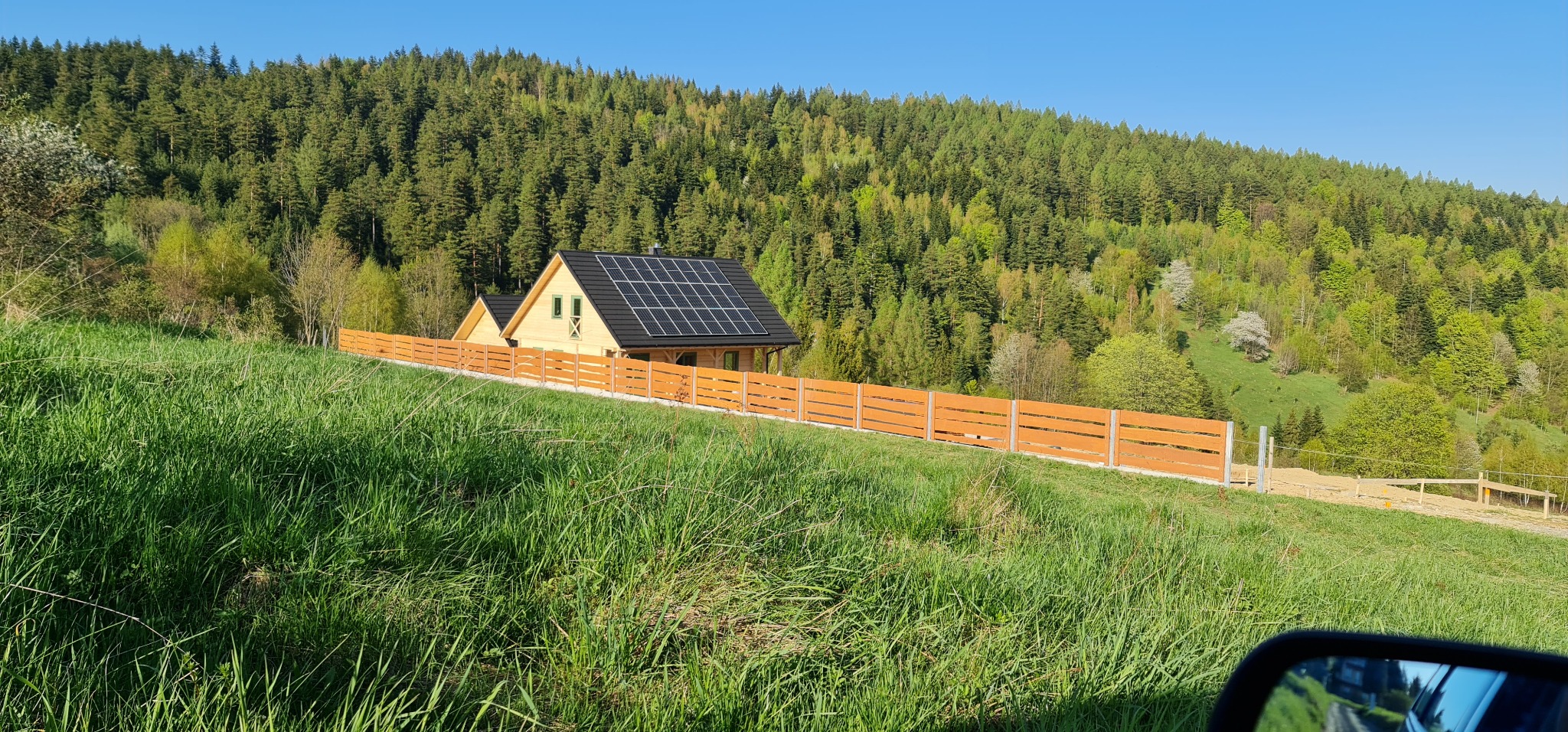 Nowoczesny dom z panelami słonecznymi na dachu, otoczony drewnianym ogrodzeniem, na tle zalesionego wzgórza w słoneczny dzień.
