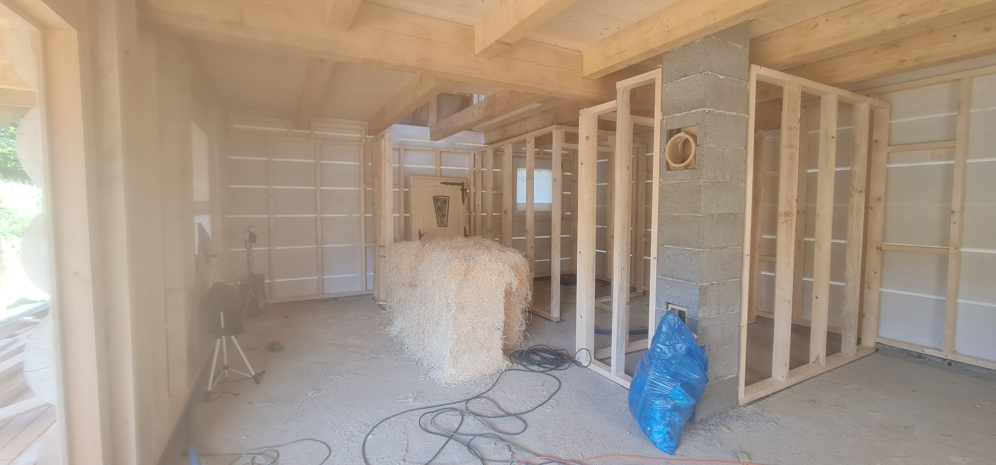 Wnętrze w trakcie budowy: drewniana konstrukcja szkieletowa ścian, izolacja z wełny drzewnej, komin z bloczków betonowych z otworem wentylacyjnym, niebieski worek na odpady budowlane.