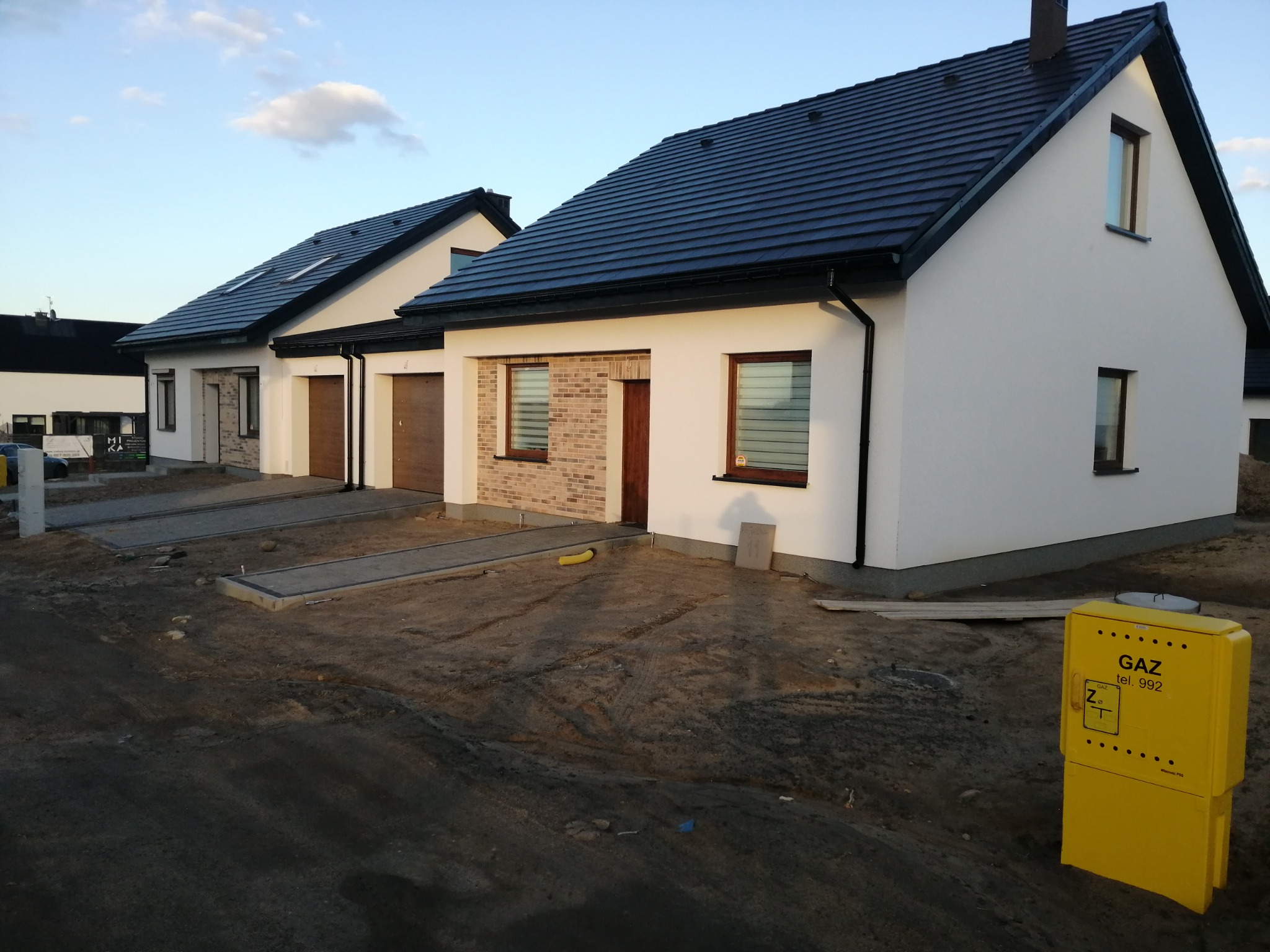 Dwa identyczne, nowo wybudowane domy jednorodzinne z białymi elewacjami i ciemną dachówką, z elementami dekoracyjnymi z cegły klinkierowej, widok z zewnątrz na jeszcze niezagospodarowany teren...