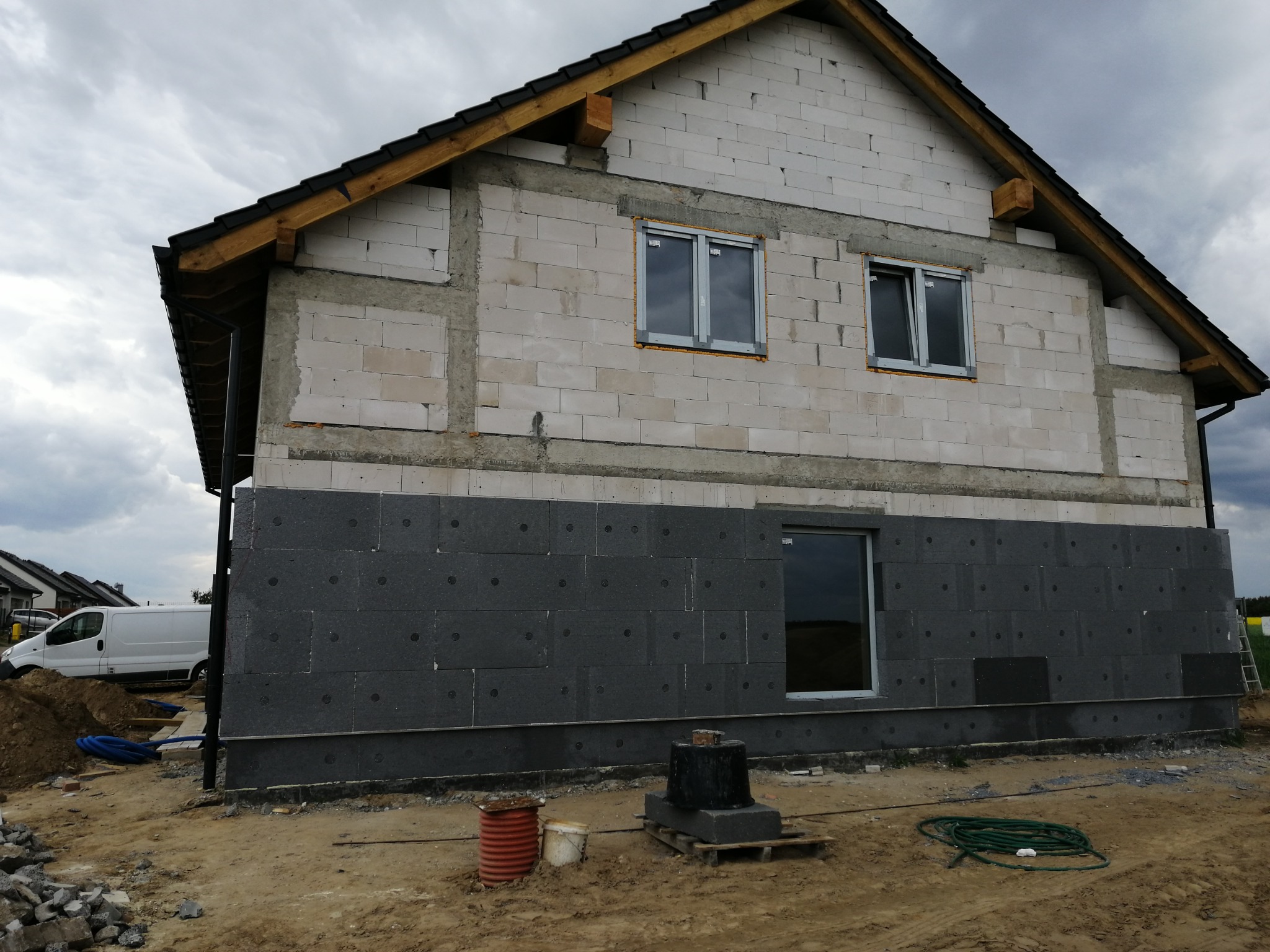 Dom w trakcie ocieplania: szare płyty izolacyjne na dolnej części elewacji, białe bloczki na górze, zamontowane okna, widoczny teren budowy i biały samochód dostawczy w tle.