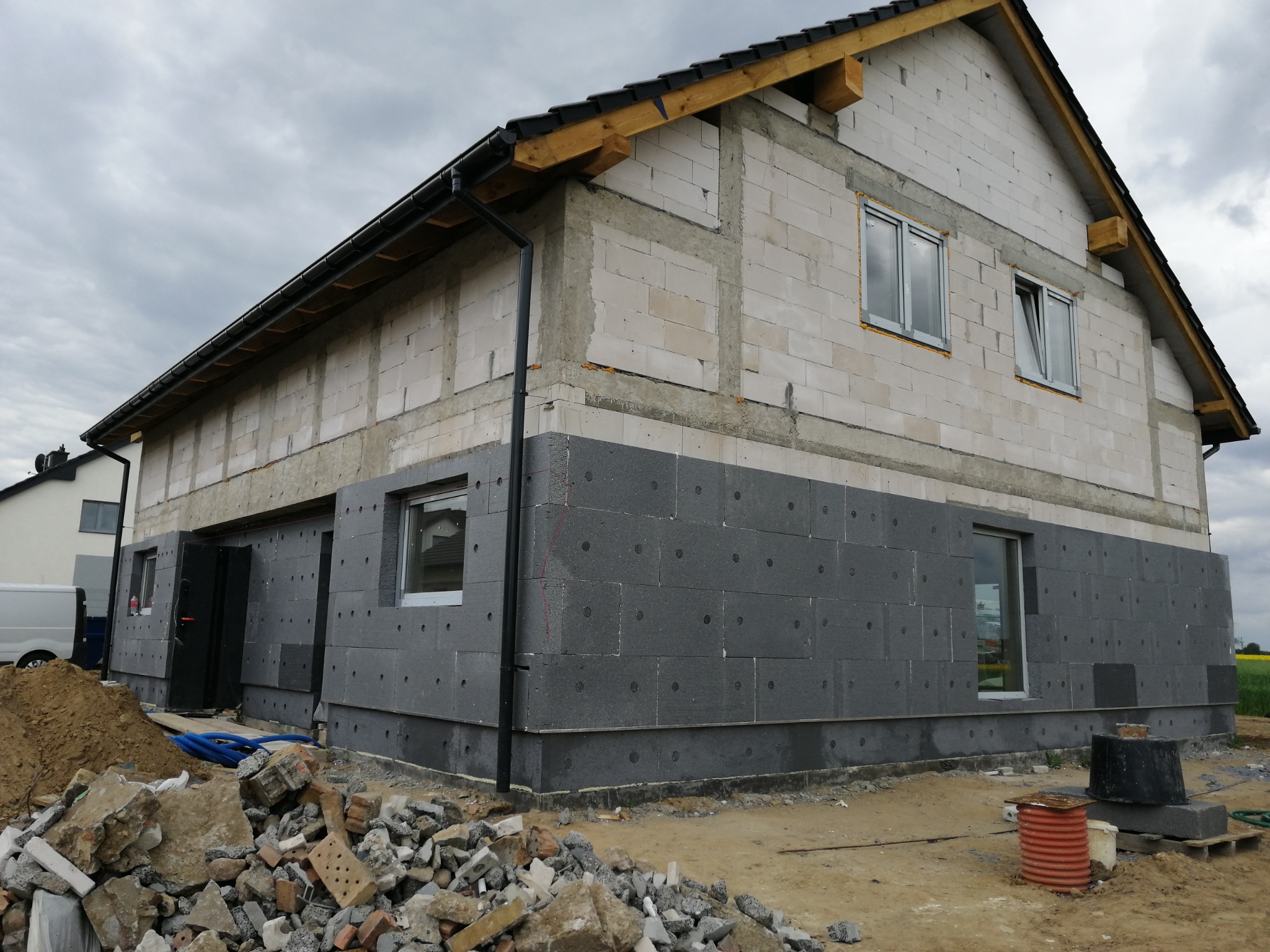 Dwukondygnacyjny budynek w trakcie ocieplania, z widocznymi szarymi płytami izolacyjnymi na dolnej części elewacji, niezabezpieczone okna i gruz budowlany przed budynkiem.