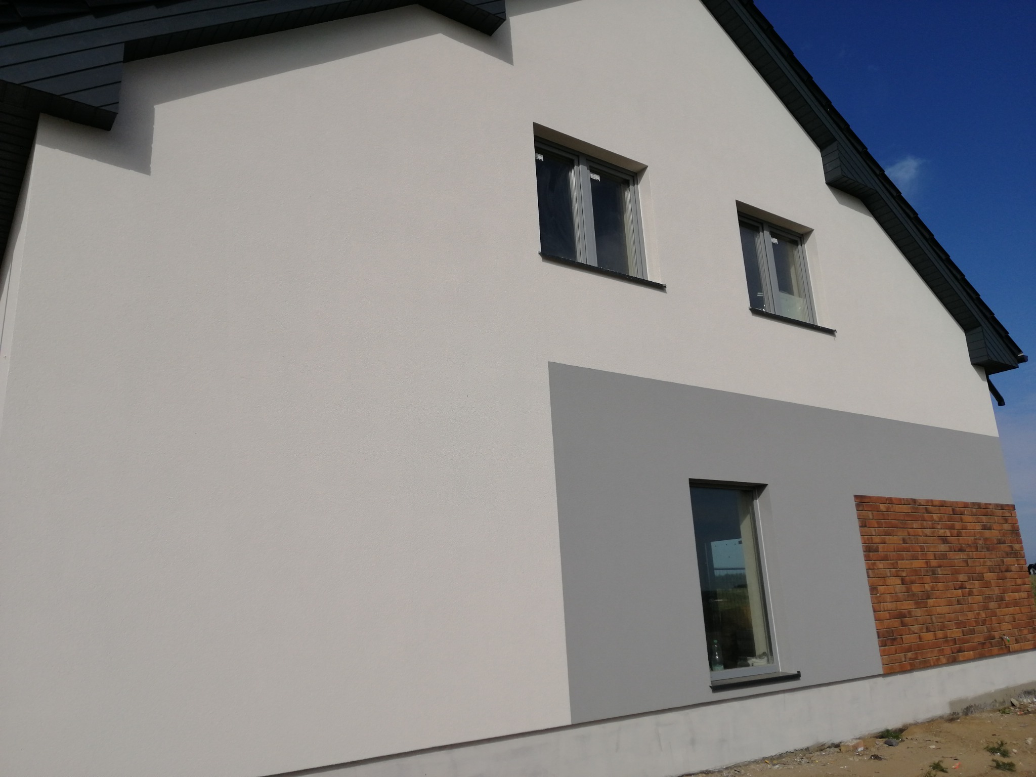 Nowoczesna elewacja domu z białym tynkiem, szarym akcentem i fragmentem ściany wykończonym cegłą klinkierową, okna z szarymi ramami.