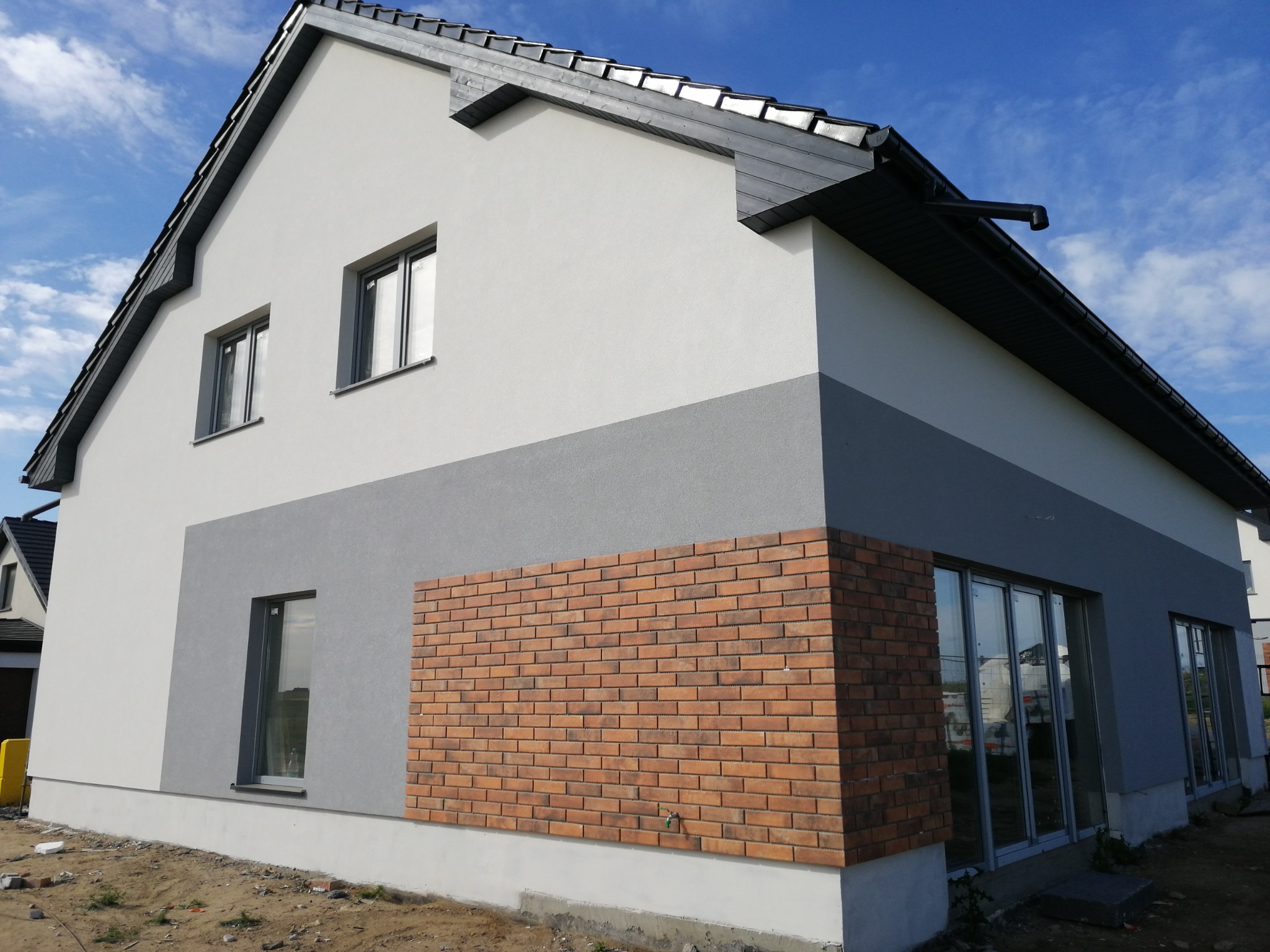 Nowoczesny dom jednorodzinny z dwukolorową elewacją: biała górna część i szara dolna, z fragmentem ściany wykończonym czerwoną cegłą klinkierową, widoczny fragment dachu z szarą dachówką i rynnami,...