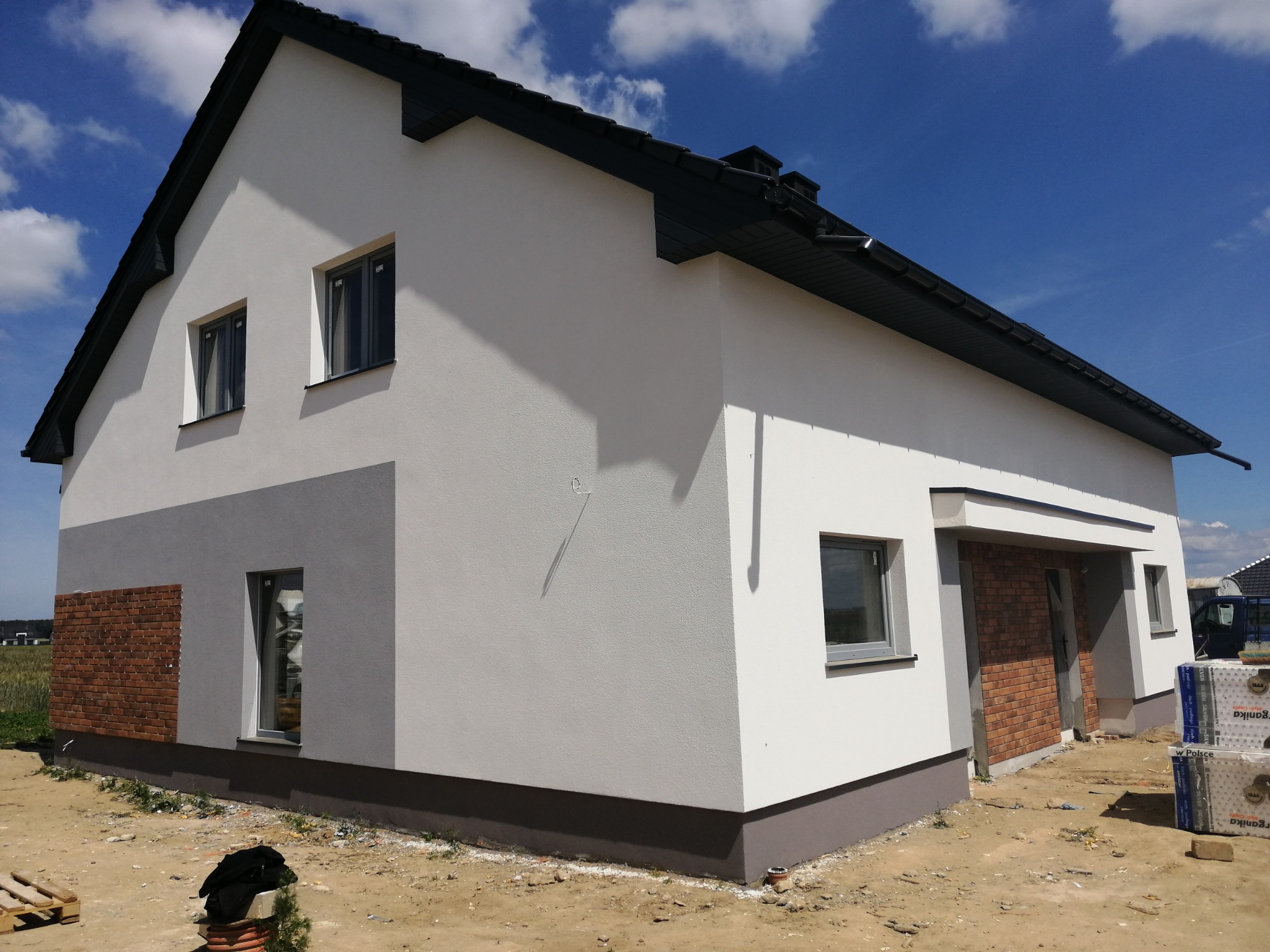 Nowoczesny dom jednorodzinny z białą elewacją, szarymi akcentami i fragmentami wykończonymi cegłą, widoczne materiały budowlane wokół.