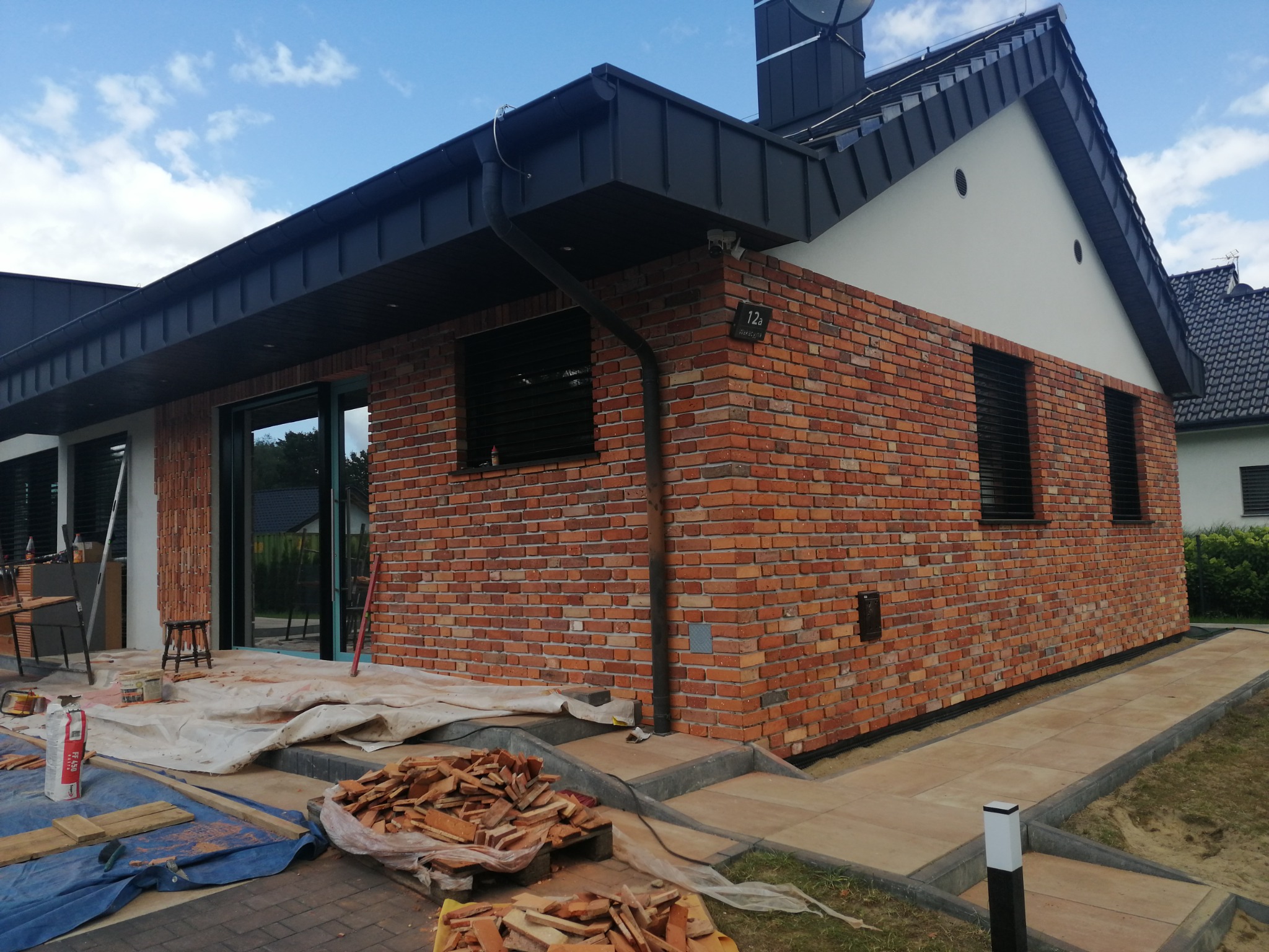 Fragment domu jednorodzinnego z nową elewacją z cegły klinkierowej, widoczne pozostałości materiału i narzędzia pracy, na ścianie numer posesji 12a.