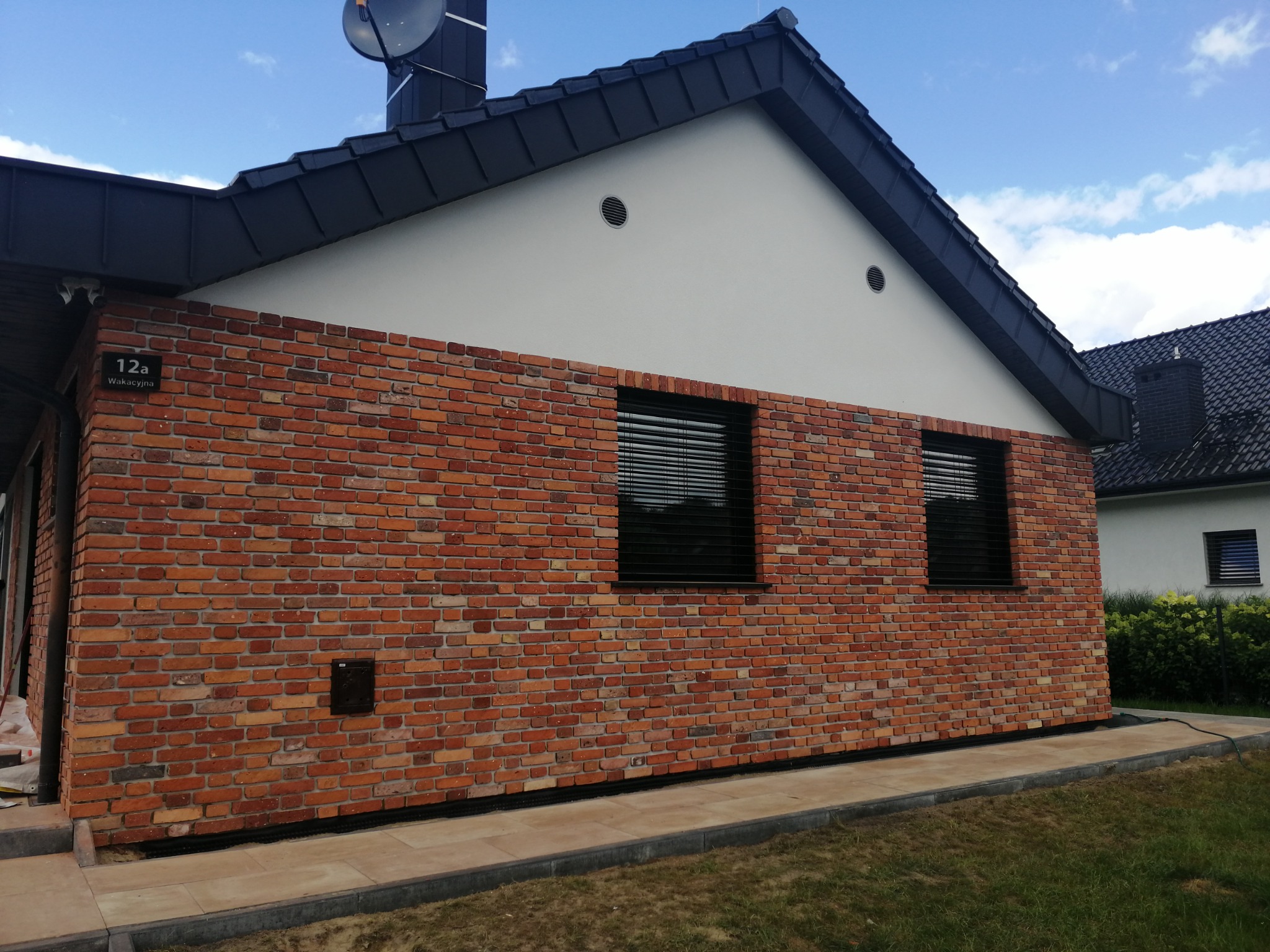 Fragment elewacji budynku z cegły klinkierowej w odcieniach czerwieni i pomarańczy, widoczne dwa okna z żaluzjami, numer domu 12a oraz fragment dachu z anteną satelitarną.