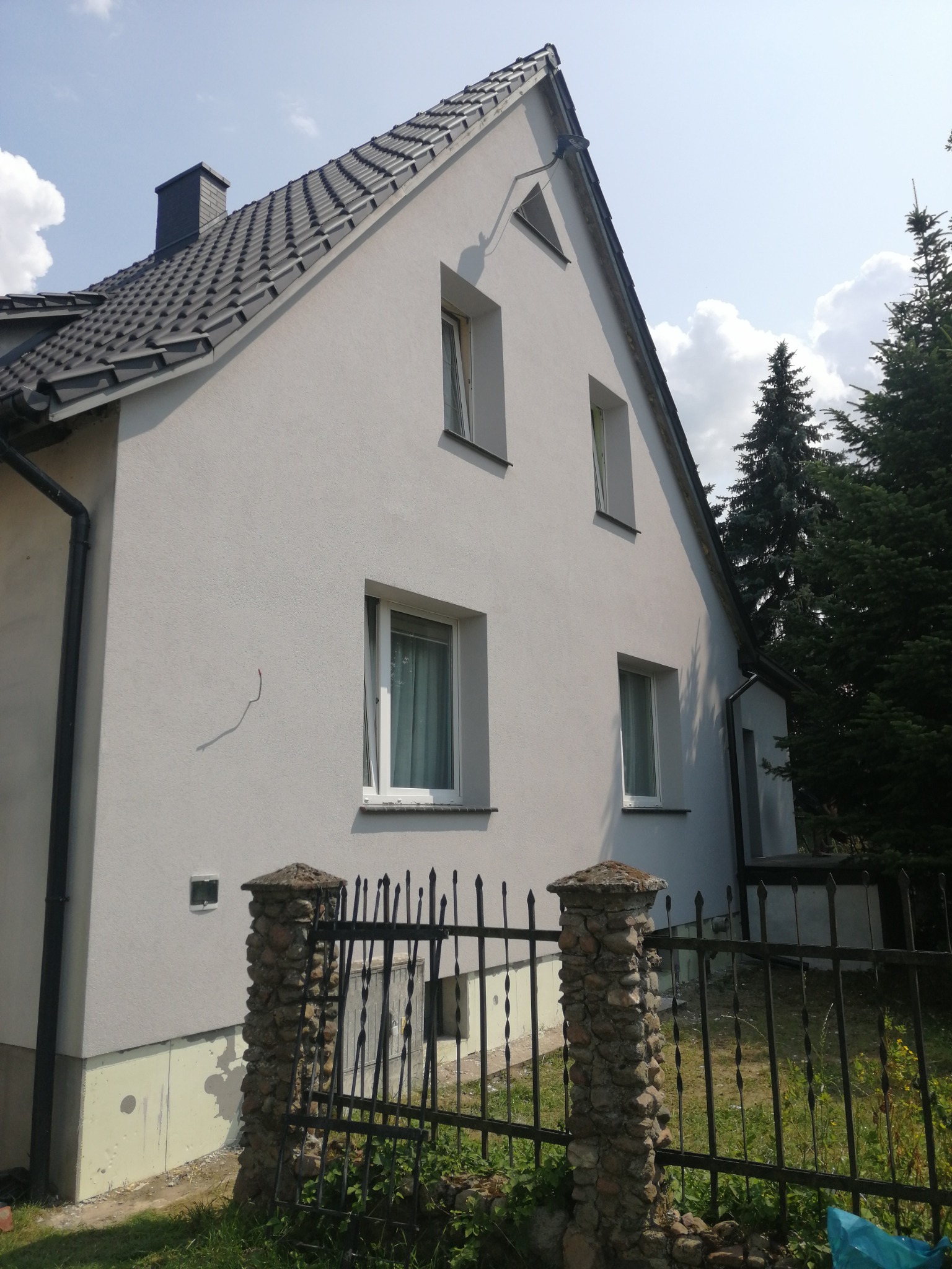 Jasnoszary dom jednorodzinny z ciemną dachówką, widok z boku, z metalowym ogrodzeniem i kamiennymi słupkami na pierwszym planie.