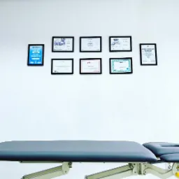 Szary stół rehabilitacyjny z regulowaną wysokością na tle białej ściany z powieszonymi certyfikatami i dyplomami w czarnych ramkach.