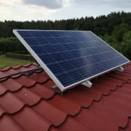 Panel fotowoltaiczny zamontowany na czerwonym dachu z dachówki ceramicznej, widoczne elementy montażowe i przewody, w tle drzewa i częściowo zachmurzone niebo.