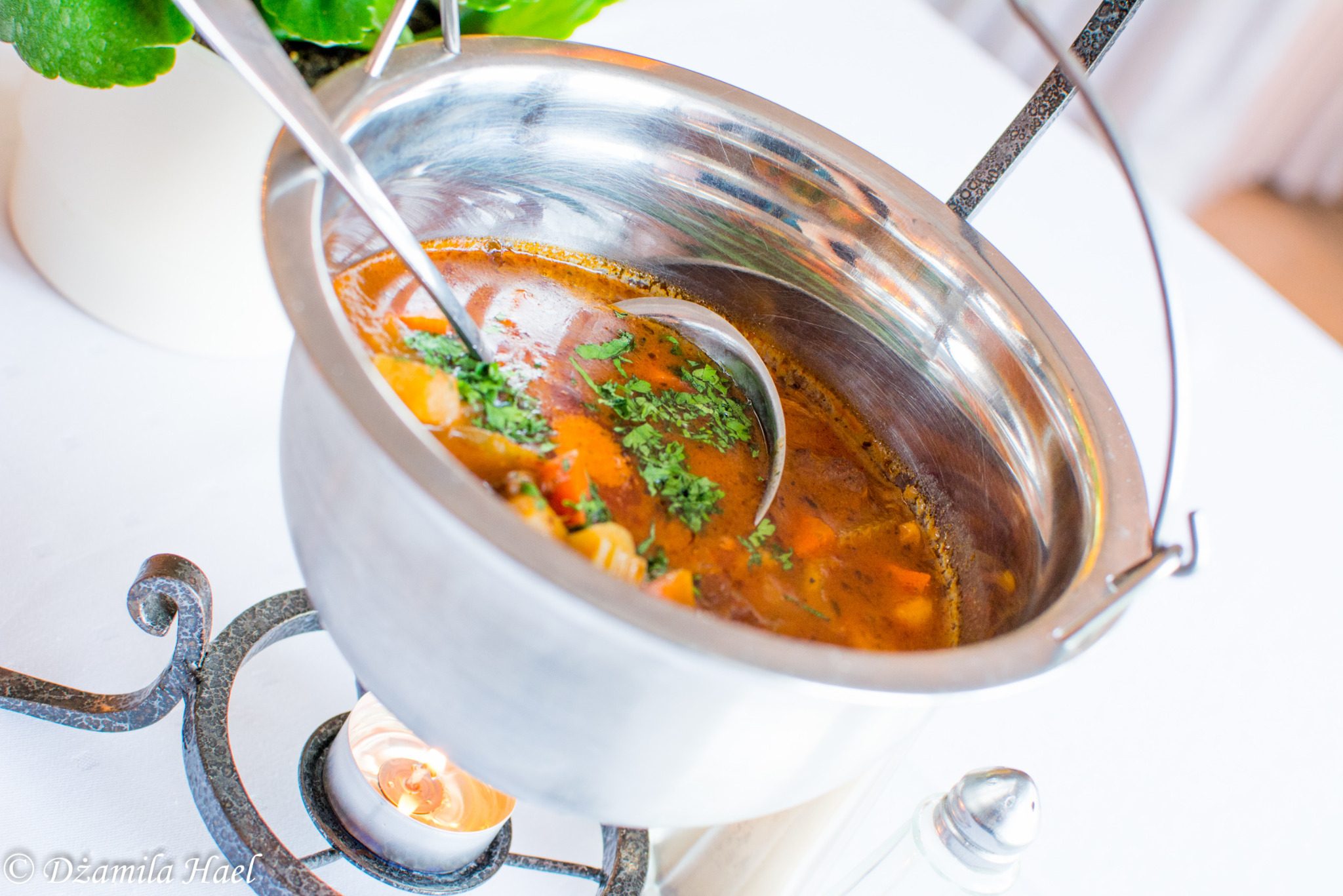 Metalowy kociołek z zupą gulaszową, doprawioną świeżą zieloną pietruszką, zawieszony nad podgrzewaczem z palącą się świeczką, na białym obrusie.