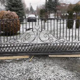 Kuta, metalowa brama wjazdowa z ozdobnymi elementami na tle zaśnieżonego ogrodu i domu w pochmurny dzień.