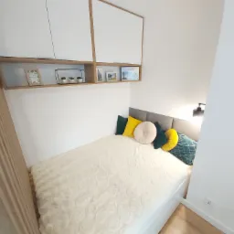 Mała sypialnia z łóżkiem, wiszącymi szafkami i półką z dekoracjami, utrzymana w jasnej kolorystyce.