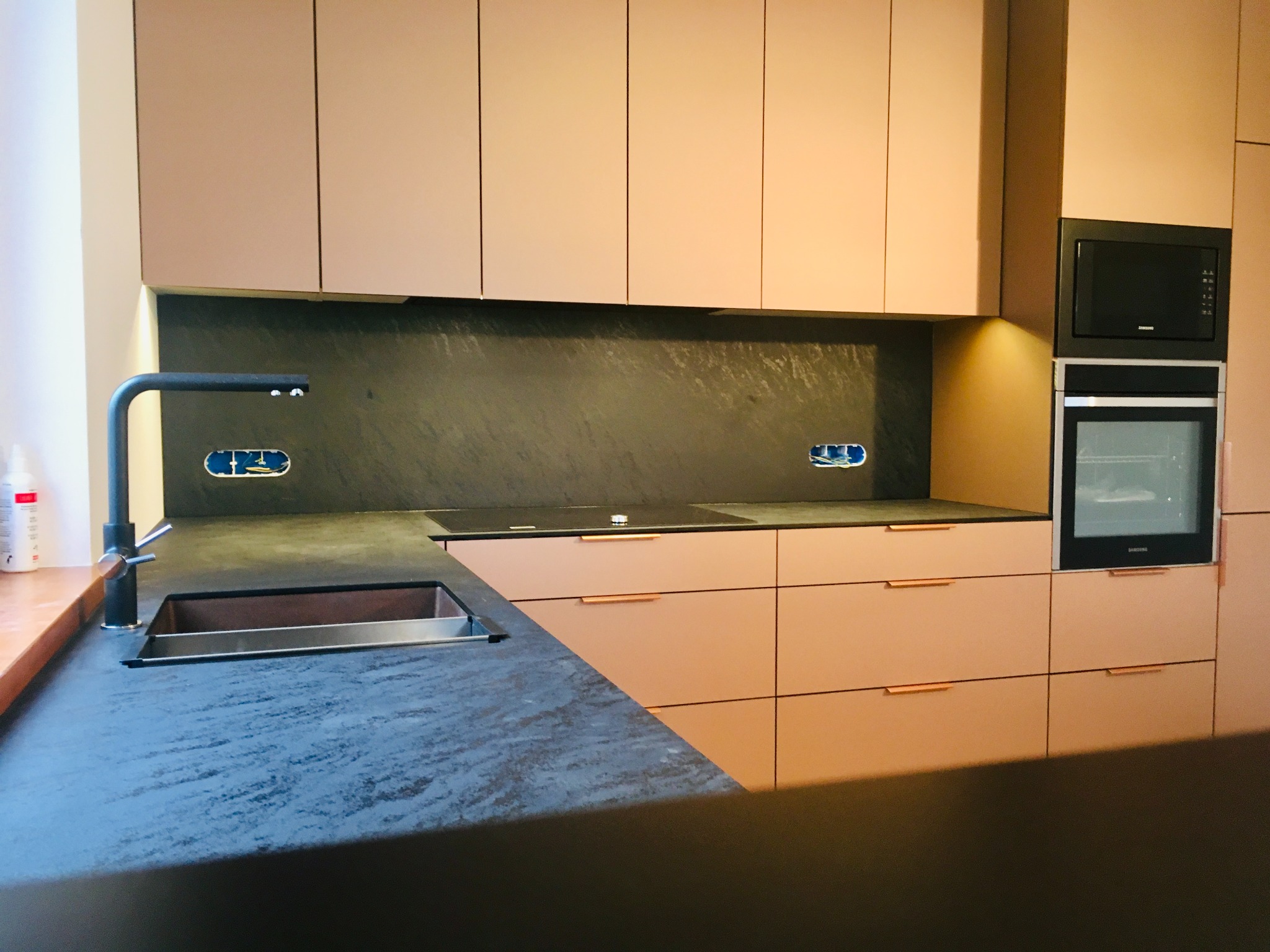 Nowoczesna kuchnia z frontami w odcieniach pudrowego różu, czarnym blatem z delikatną fakturą i minimalistycznymi, miedzianymi uchwytami. Widoczne gniazdka elektryczne nad blatem, czarny zlew...