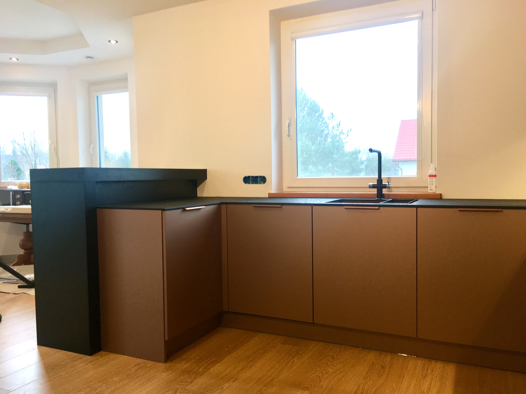 Nowoczesna kuchnia z brązowymi frontami szafek, czarnym blatem i armaturą. Widoczne gniazdko elektryczne nad blatem i fragment stołu w tle.