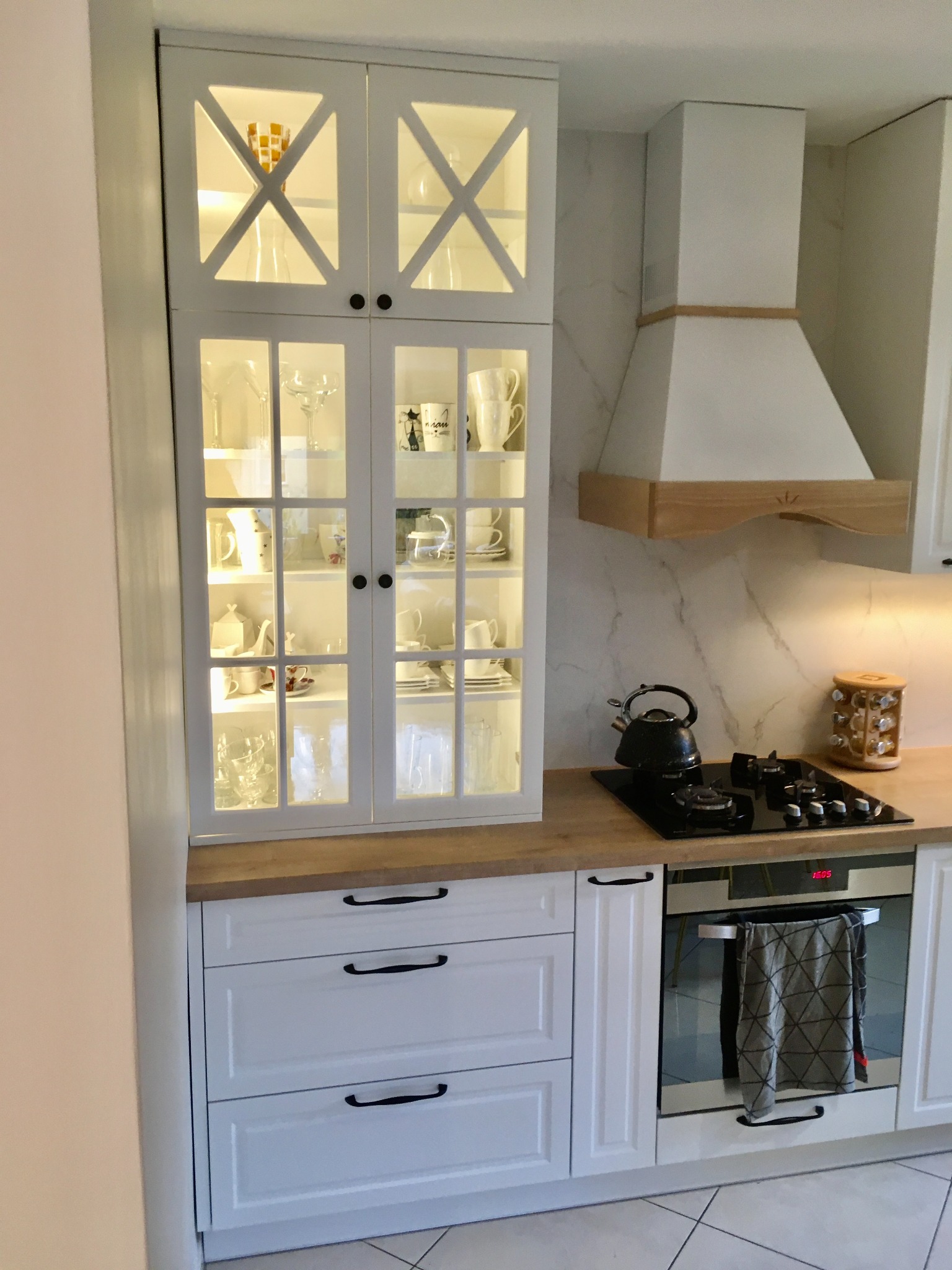 Biała zabudowa kuchenna z przeszkloną witryną, czajnikiem na płycie grzewczej i okapem z drewnianymi akcentami.