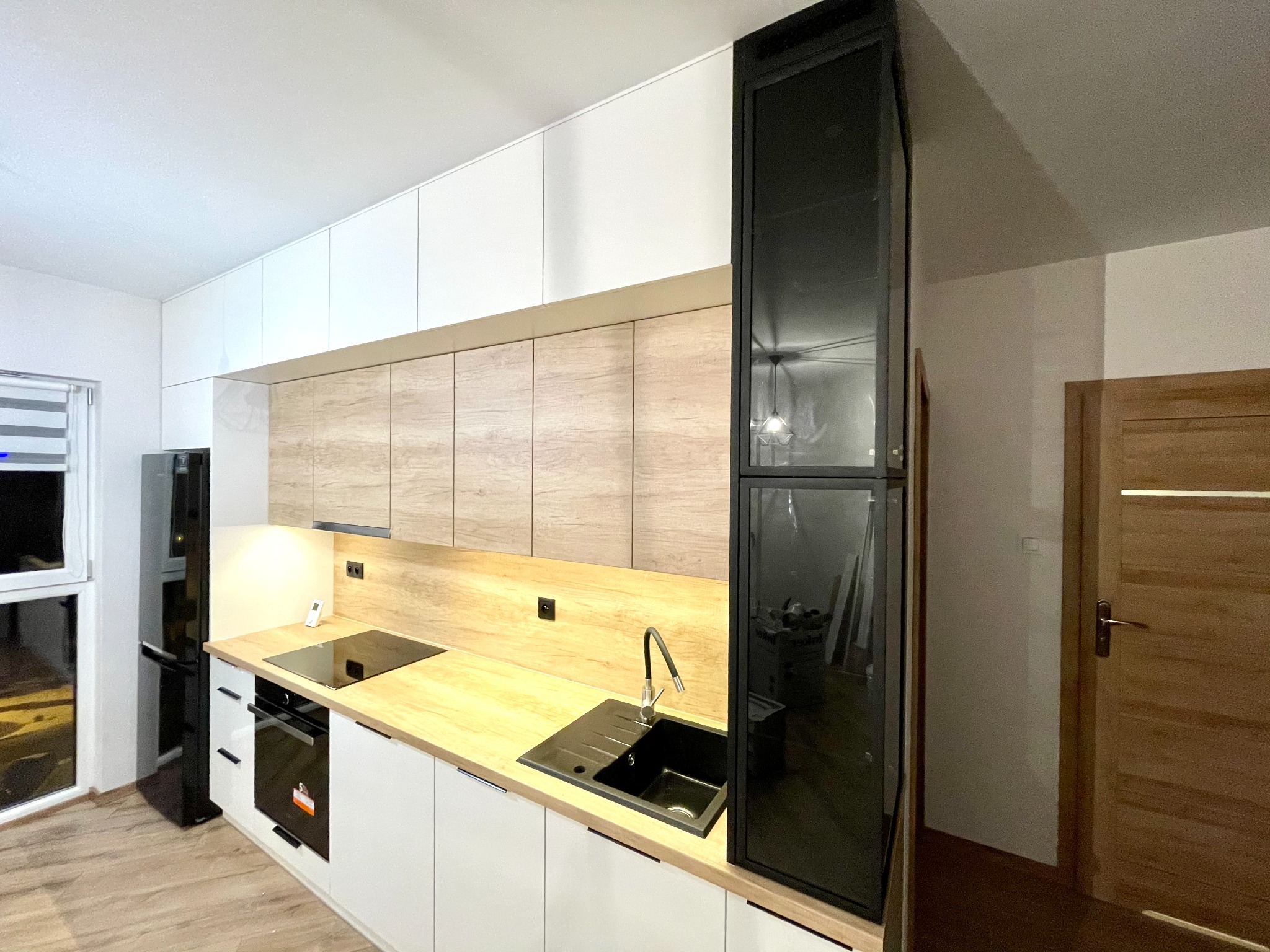 Nowoczesna kuchnia z zabudową w odcieniach bieli i drewna w Brzostku, z czarnym zlewem i płytą indukcyjną, widok na lodówkę i drzwi.