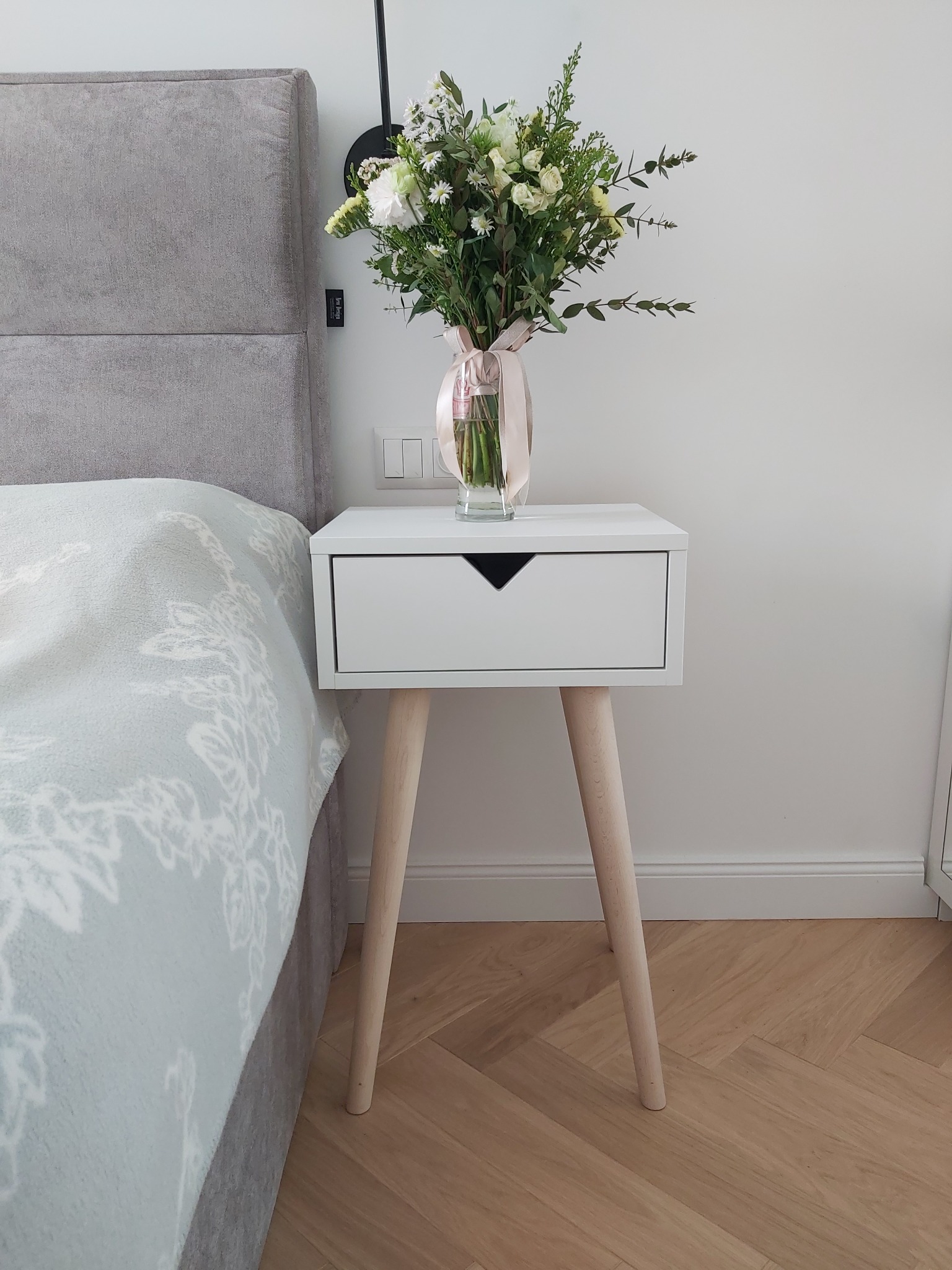 Biała szafka nocna z szufladą i drewnianymi nóżkami, na której stoi wazon z bukietem kwiatów, obok łóżko z szarym wezgłowiem i wzorzystą narzutą, parkiet ułożony w jodełkę.