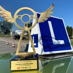 Nauka jazdy doszkalanie Grójec - Nagroda 'Instruktor Jazdy Roku 2025' z symbolem kierownicy ze skrzydłami, obok znak 'L' na dachu auta. W tle droga i drzewa. Słoneczny dzień.