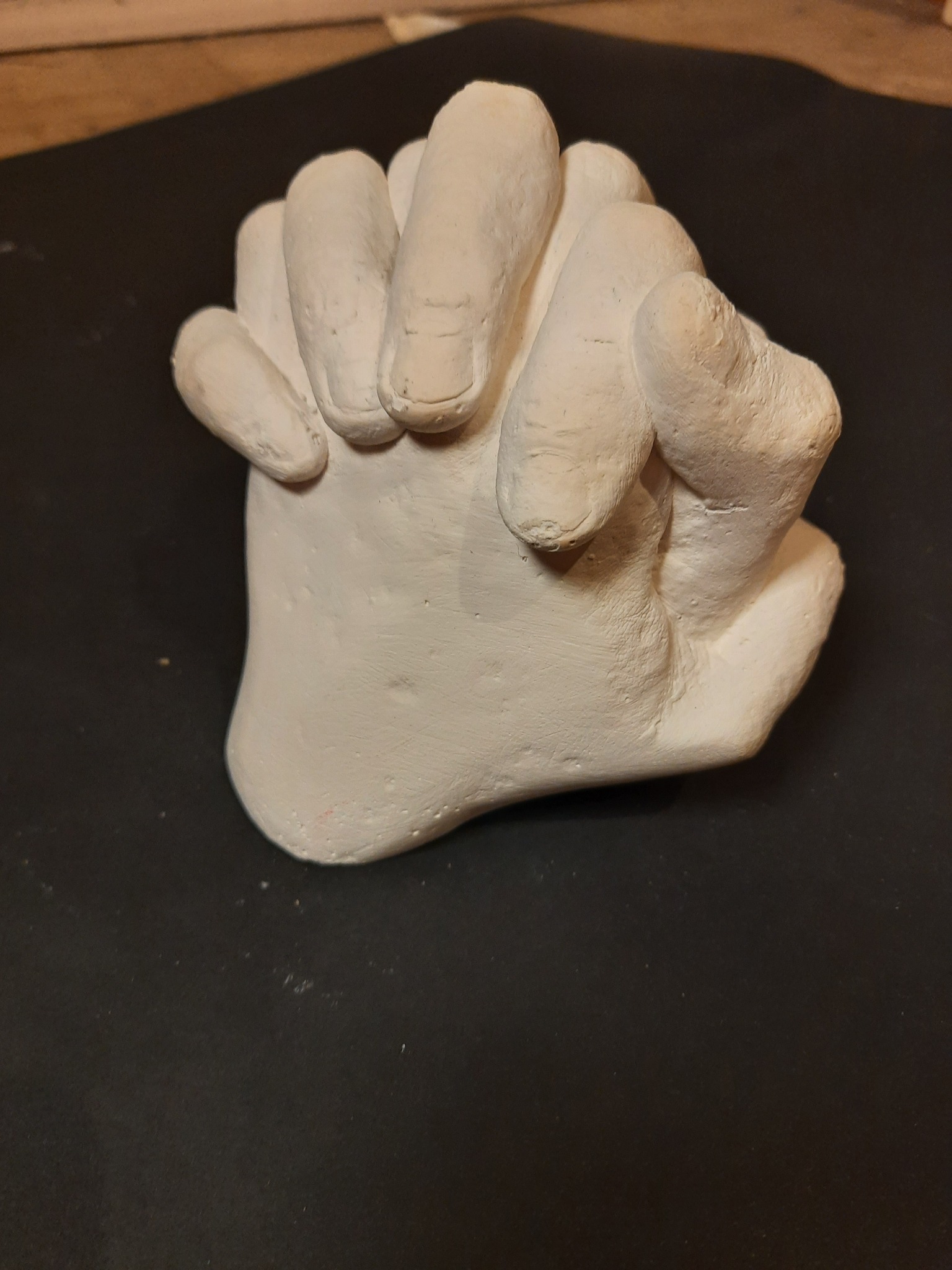 Biały odlew gipsowy złączonych dłoni na ciemnym tle, ukazujący detale faktury i nierówności materiału.