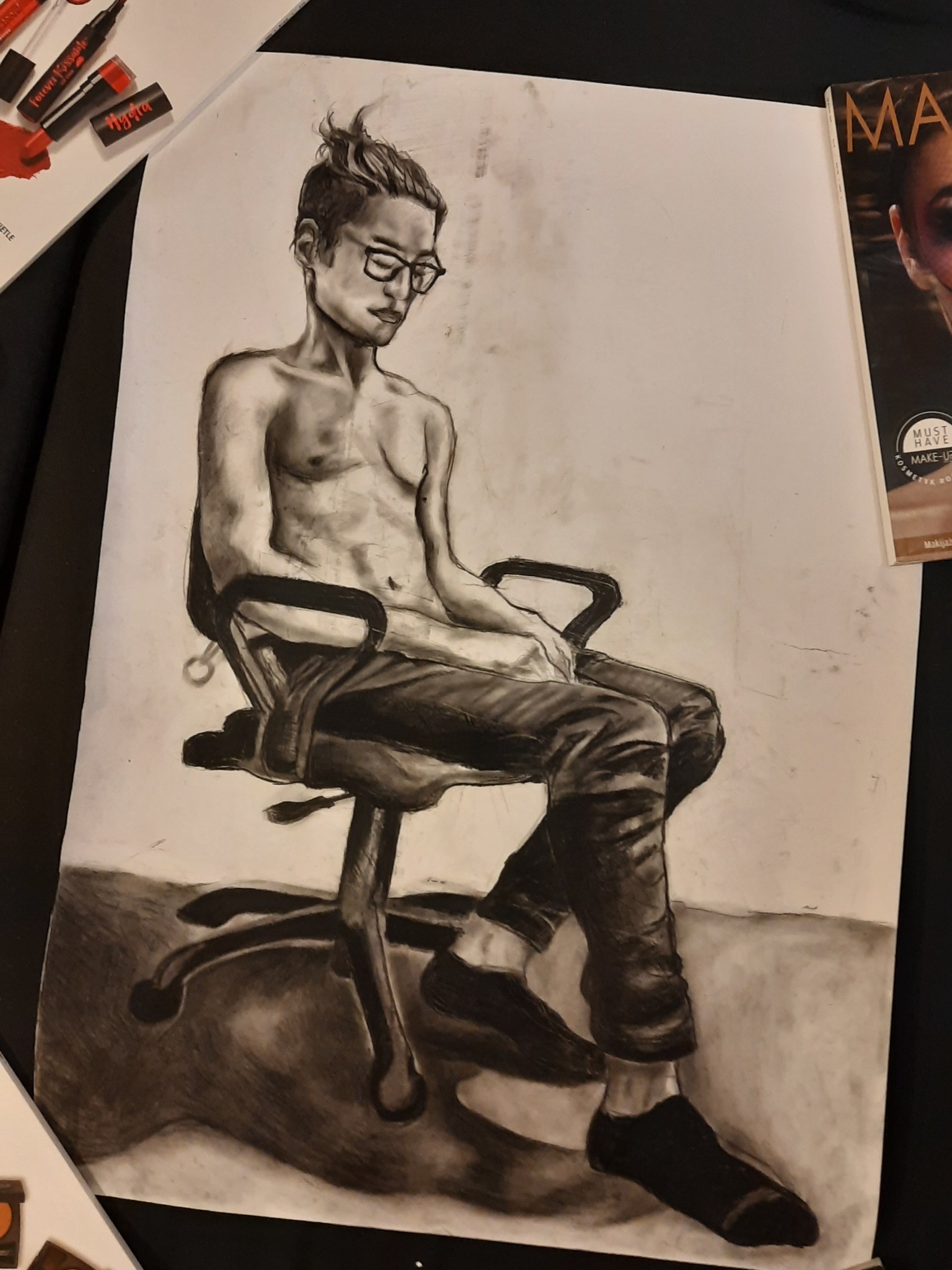 Ręcznie rysowany węglem portret mężczyzny w okularach siedzącego na krześle biurowym, z magazynem kosmetycznym i kosmetykami w tle.