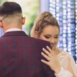 Para młoda w tańcu: panna młoda w białej sukni z koronkowymi rękawami obejmuje pana młodego w bordowym garniturze w kratę, tło rozświetlone dekoracyjnymi lampkami.