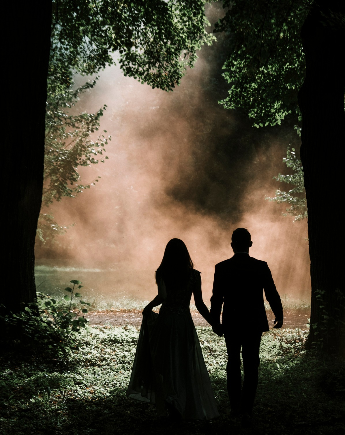 Sylwetki pary trzymającej się za ręce, idącej ścieżką w lesie, z efektem dymu w tle, tworzącym romantyczną atmosferę.