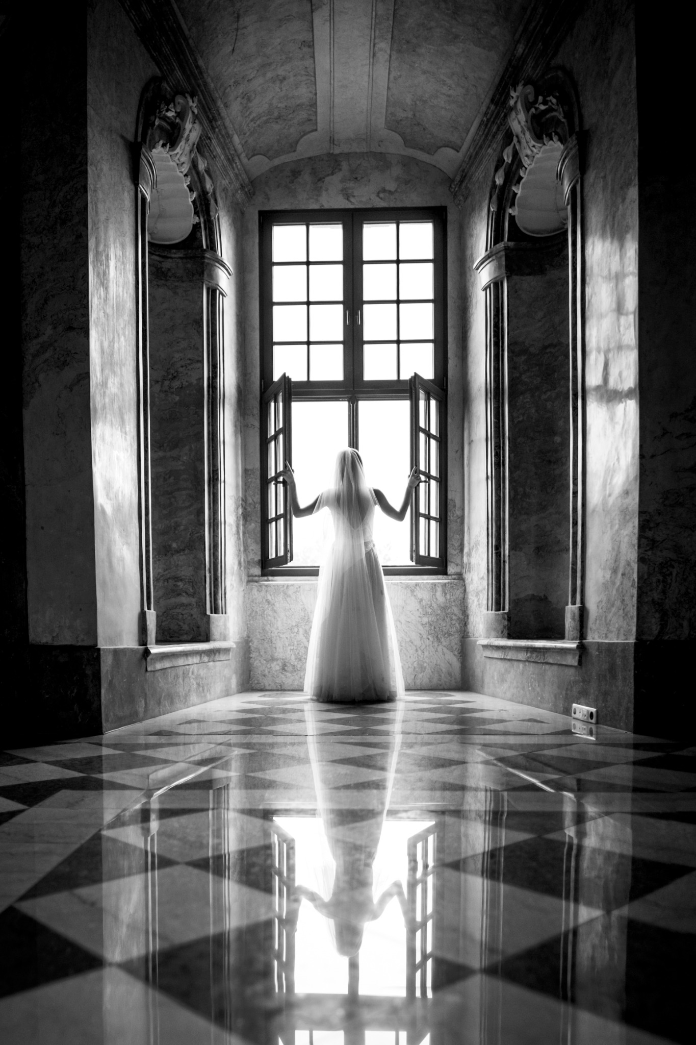 Czarno-białe zdjęcie panny młodej w sukni i welonie, stojącej tyłem przy otwartym oknie w marmurowym pomieszczeniu z szachownicową podłogą, której odbicie widoczne jest w podłodze.