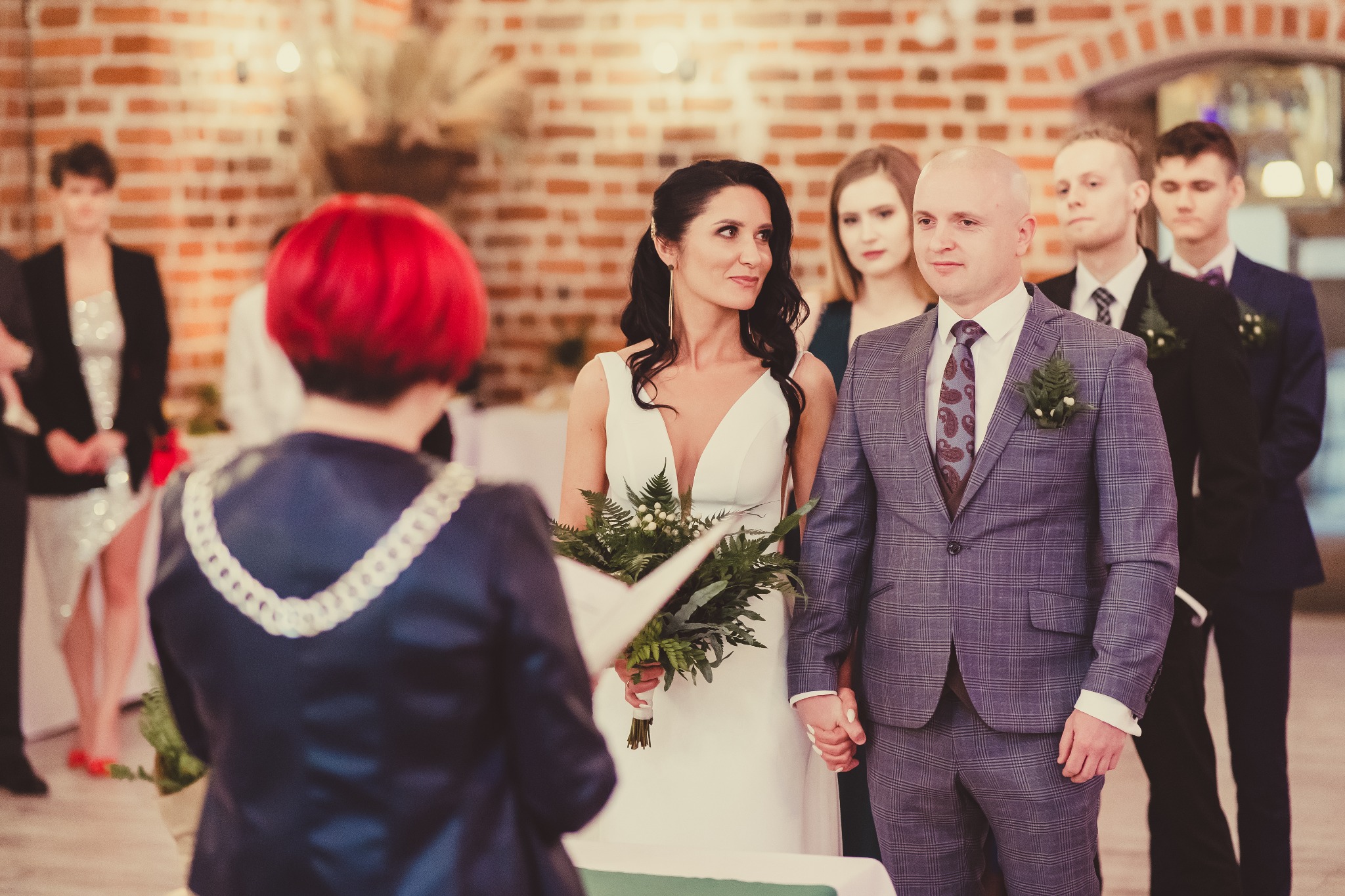 Para młoda podczas ceremonii ślubnej w ceglanym wnętrzu, panna młoda w białej sukni trzyma bukiet z zielonych liści, pan młody w szarym garniturze w kratę, w tle świadkowie i osoba prowadząca...