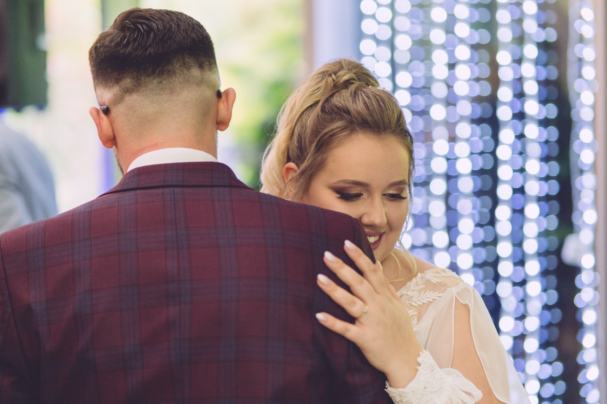 Para młoda w tańcu: panna młoda w białej sukni z koronkowymi rękawami obejmuje pana młodego w bordowym garniturze w kratę, tło rozświetlone dekoracyjnymi lampkami.