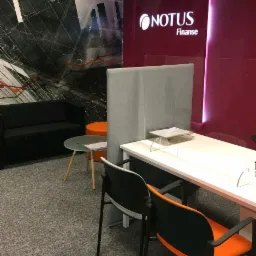 Wnętrze biura finansowego z logo Notus Finanse, biurko z przegrodą, krzesła z pomarańczowymi siedziskami, sofa i stolik kawowy na tle abstrakcyjnej grafiki ściennej.