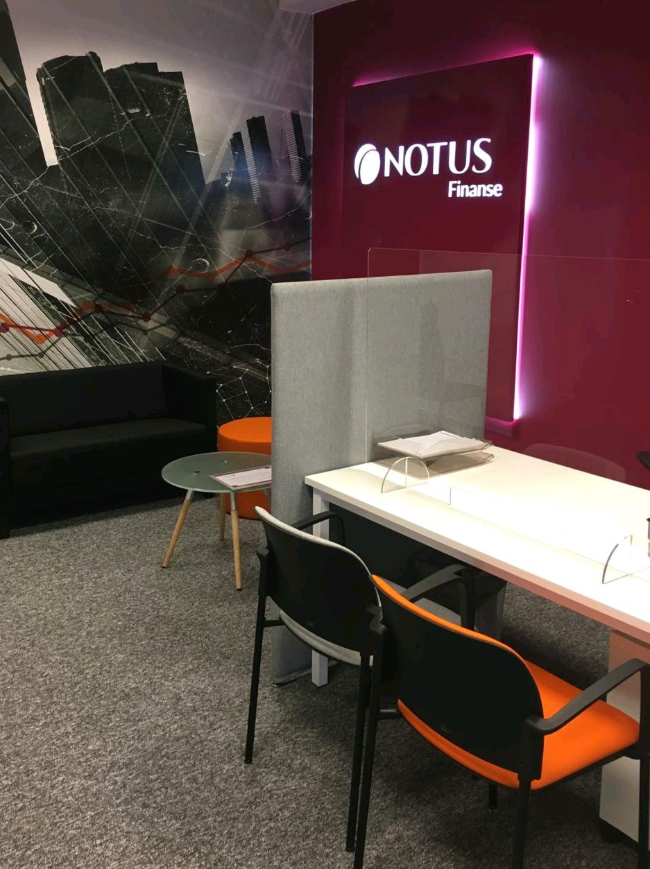 Wnętrze biura finansowego z logo Notus Finanse, biurko z przegrodą, krzesła z pomarańczowymi siedziskami, sofa i stolik kawowy na tle abstrakcyjnej grafiki ściennej.