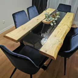 Stół z blatem z drewna o nieregularnych krawędziach i szklanym środkiem, otoczony sześcioma ciemnoniebieskimi, tapicerowanymi krzesłami na czarnych nogach, na podłodze z paneli.