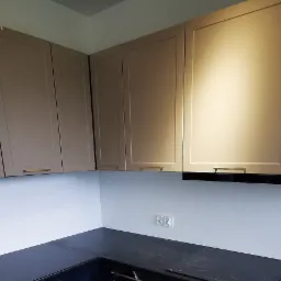 Górne szafki kuchenne w odcieniach beżu z minimalistycznymi uchwytami, zamontowane nad czarnym blatem i płytą indukcyjną, widoczne gniazdko elektryczne na ścianie.