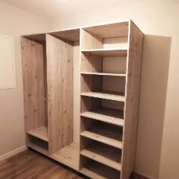 Wnętrze garderoby z jasnego drewna z otwartymi półkami i przestrzenią na wieszaki, widok na podłogę z paneli.