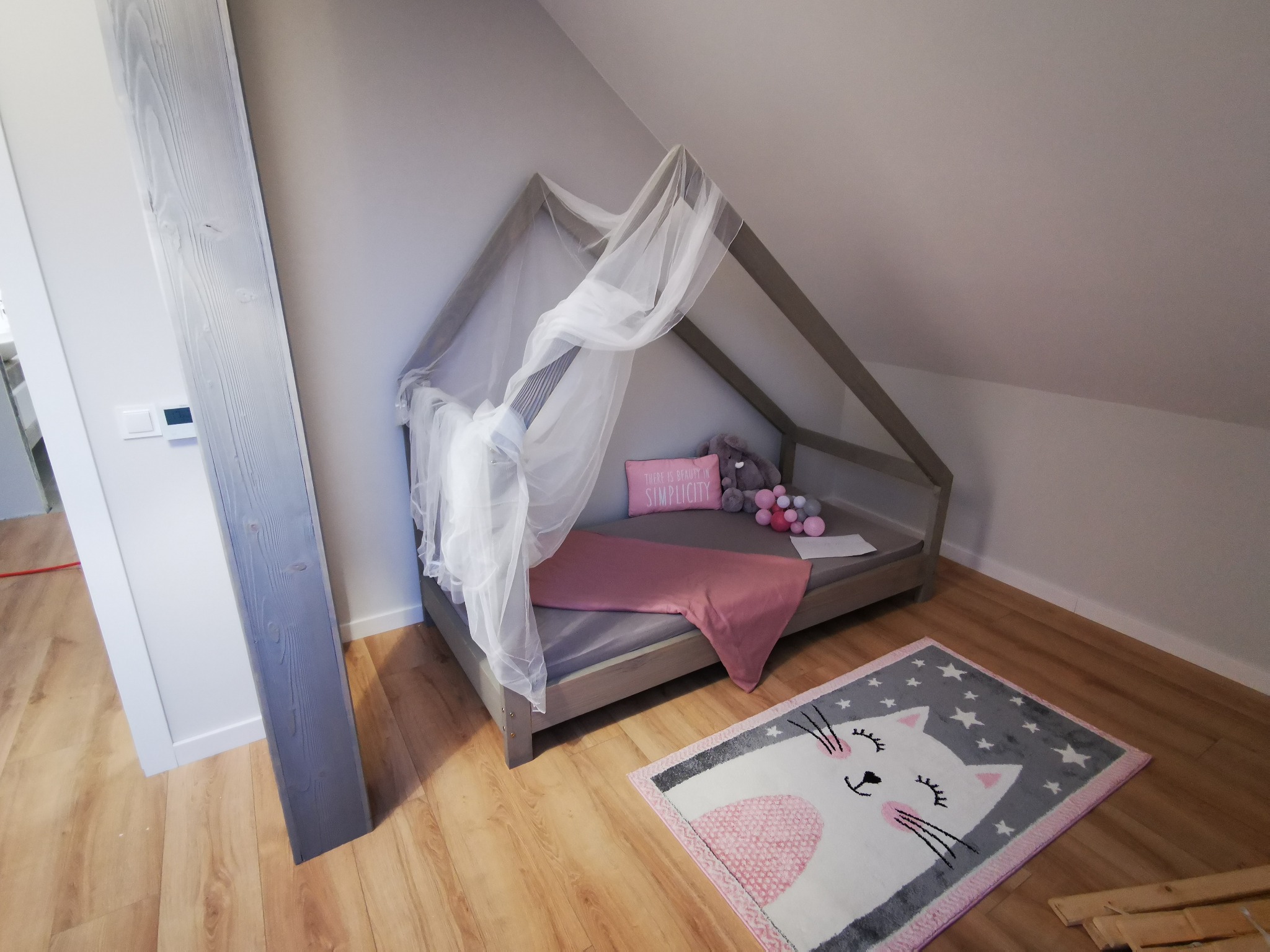 Szare łóżko dziecięce w kształcie domku z baldachimem, różowym kocykiem i poduszką z napisem 'Simplicity', na podłodze dywanik z motywem kota i gwiazdek, fragment drewnianej podłogi i szara kolumna.