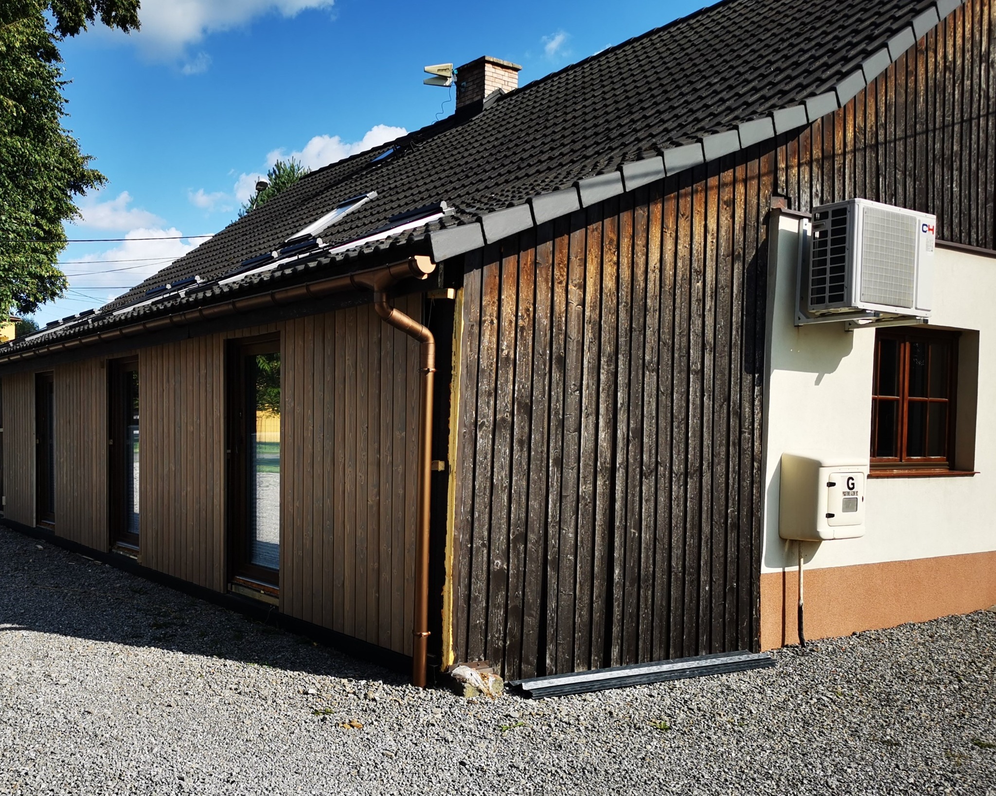 Nowoczesny dom z drewnianą elewacją częściowo w kolorze naturalnego drewna, a częściowo w ciemnym brązie, z widocznymi oknami i rynnami, na tle błękitnego nieba.