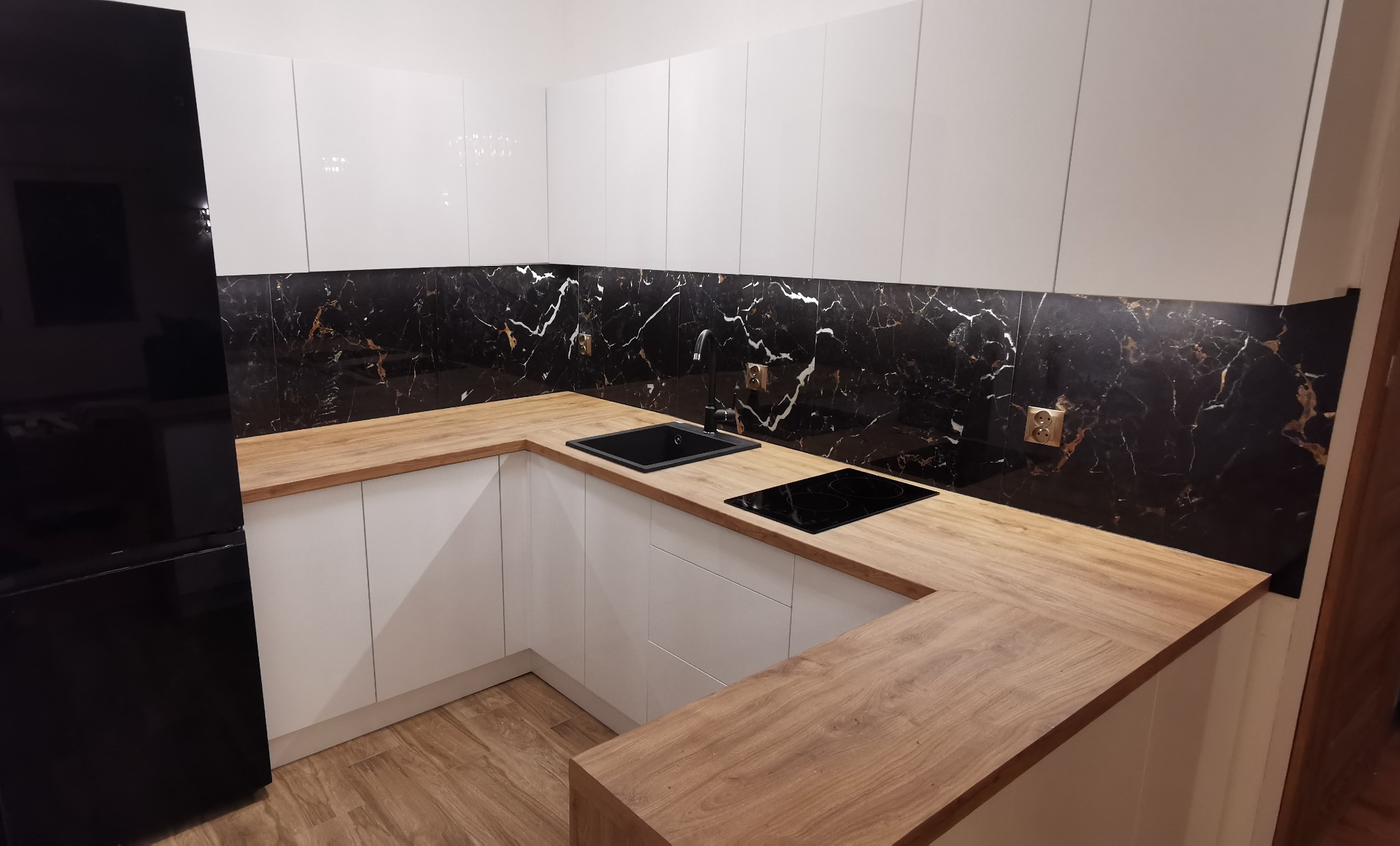 Nowoczesna kuchnia z białymi szafkami, drewnianym blatem i czarnym panelem ściennym imitującym marmur.
