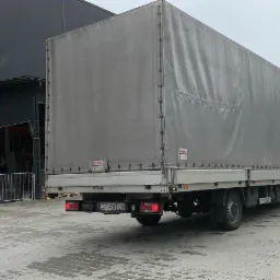 Samochód dostawczy z plandeką, zaparkowany tyłem przed halą magazynową z otwartą bramą, widoczny numer rejestracyjny CT 4618N.