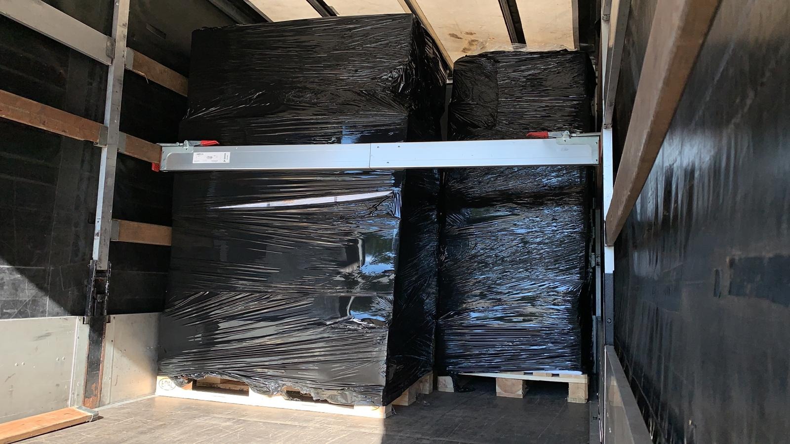 Czarne, owinięte folią ładunki na paletach, zabezpieczone w przestrzeni ładunkowej samochodu ciężarowego z aluminiową belką mocującą.