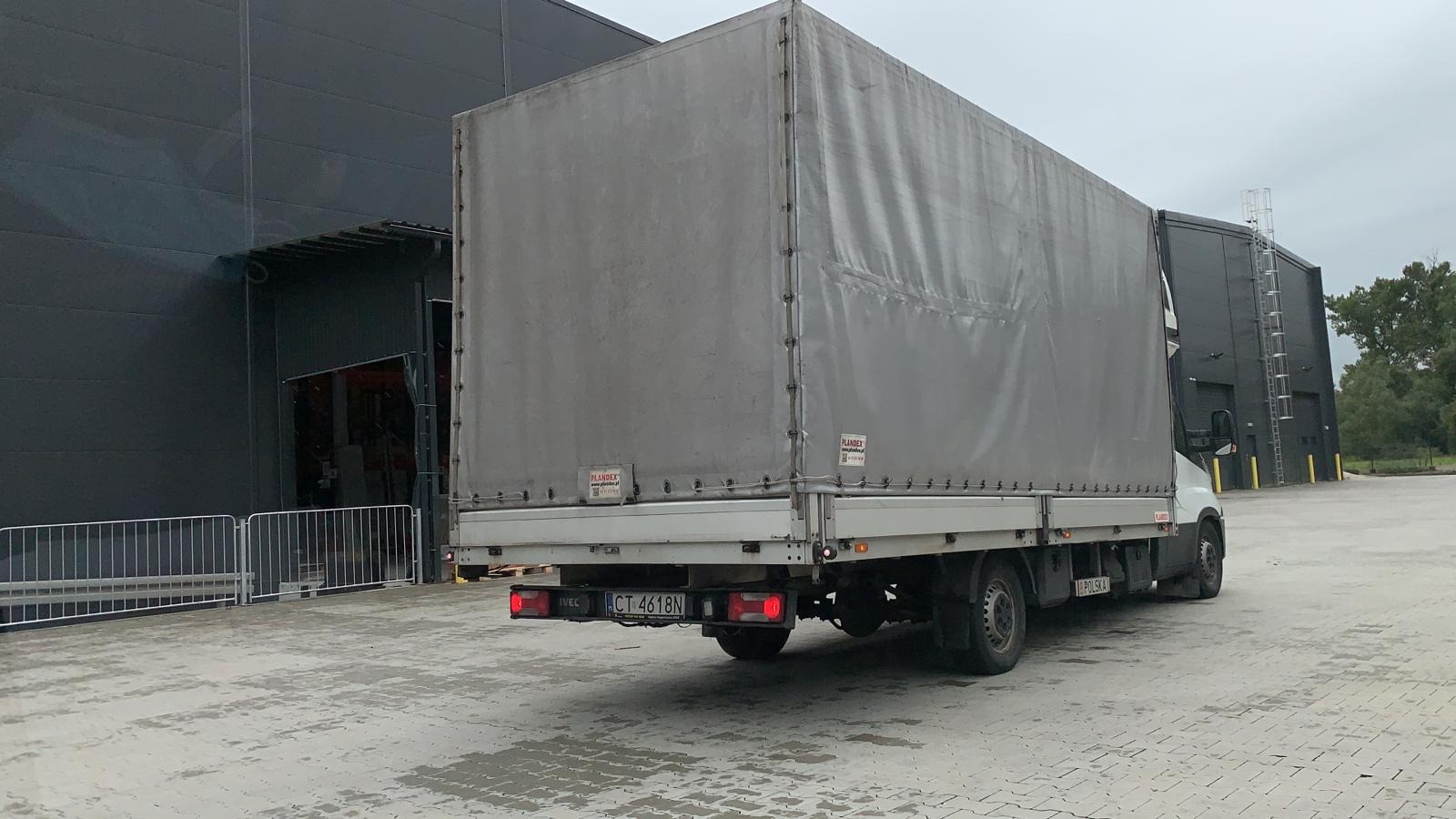 Samochód dostawczy z plandeką, zaparkowany tyłem przed halą magazynową z otwartą bramą, widoczny numer rejestracyjny CT 4618N.