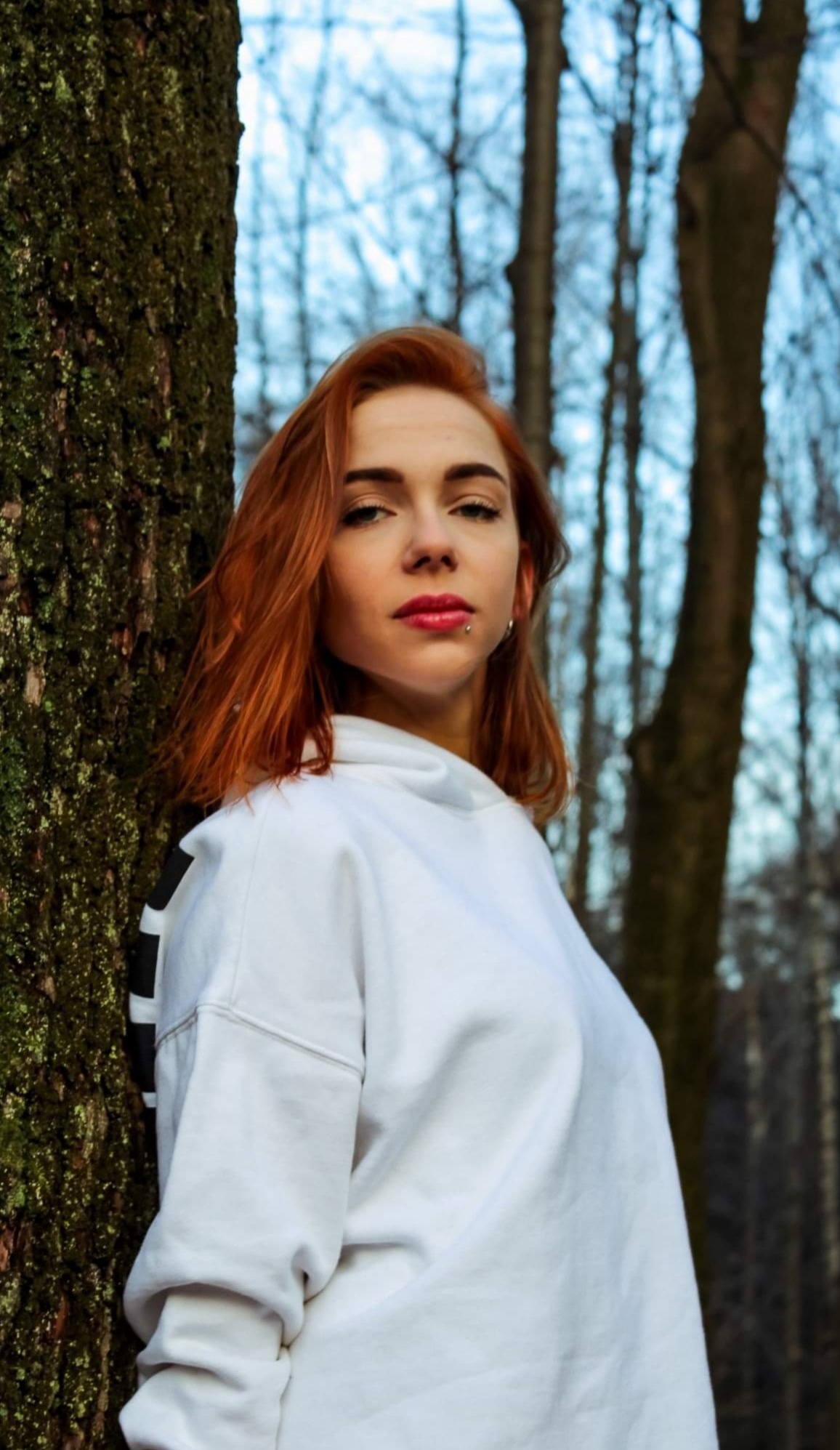 Portret rudej kobiety w białej bluzie z kapturem, opierającej się o drzewo w lesie, z piercingiem w wardze.