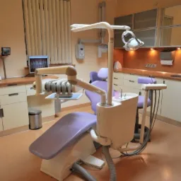 Jasne wnętrze gabinetu stomatologicznego z fotelem dentystycznym w odcieniach bieli i fioletu, widok na monitor z prześwietleniem zębów, szafki i umywalkę w tle.