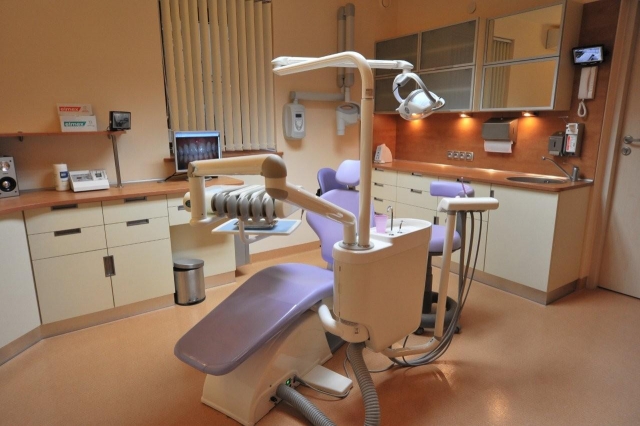 Jasne wnętrze gabinetu stomatologicznego z fotelem dentystycznym w odcieniach bieli i fioletu, widok na monitor z prześwietleniem zębów, szafki i umywalkę w tle.