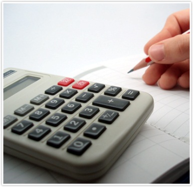 Kalkulator z szarymi przyciskami i czerwonymi akcentami leżący na notatniku w kratkę, obok dłoń trzymająca ołówek z czerwoną końcówką.