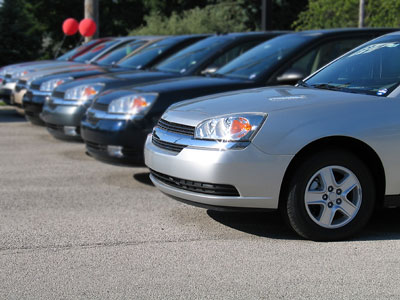 Rząd zaparkowanych samochodów osobowych różnych kolorów na placu, z czerwonymi balonami widocznymi w tle.