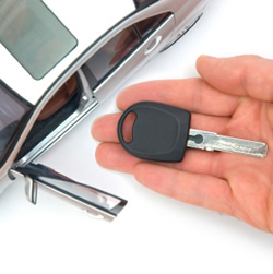 Kluczyk samochodowy na dłoni, z otwartymi drzwiami miniaturowego modelu auta w tle, na białym tle.