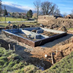 Wykop pod fundamenty domu z częściowo wylaną betonem ścianą i przygotowanymi bloczkami, widok na okolicę z pagórkami i zabudowaniami w tle.