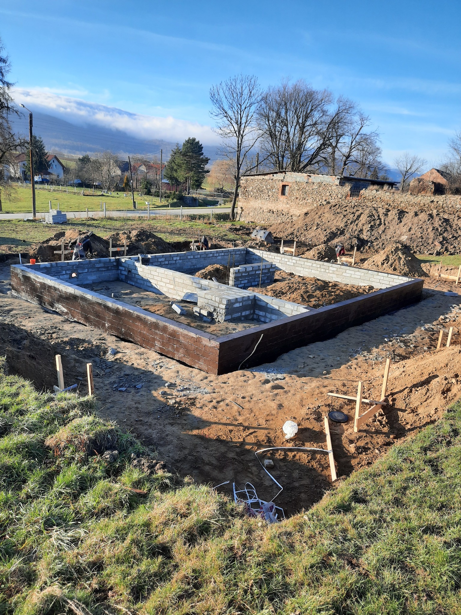 Wykop pod fundamenty domu z częściowo wylaną betonem ścianą i przygotowanymi bloczkami, widok na okolicę z pagórkami i zabudowaniami w tle.