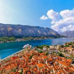 Panoramiczny widok na miasto z czerwonymi dachami, zatokę z jachtami i statkiem wycieczkowym oraz góry w tle, pod błękitnym niebem z białymi chmurami.