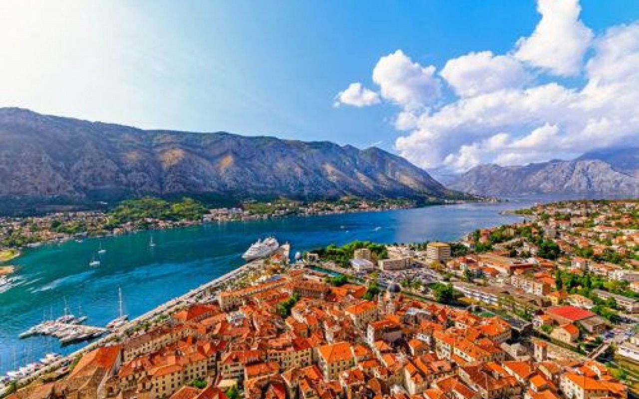 Panoramiczny widok na miasto z czerwonymi dachami, zatokę z jachtami i statkiem wycieczkowym oraz góry w tle, pod błękitnym niebem z białymi chmurami.